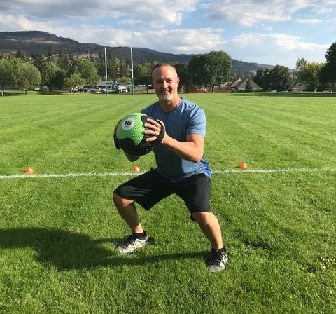 James Chicalo, fitness trainer, does a squat while holding a medicine ball, doing exercise in a green field in the South Okanagan.