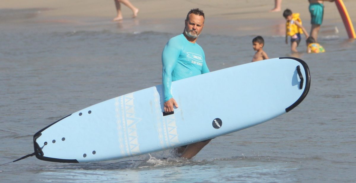 James Chicalo holds a surfboard, standing in shallow water.