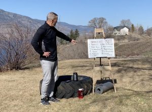 James Chicalo stands in a field overlooking Okanagan Lake, with a white board describing the HIIT workout video.